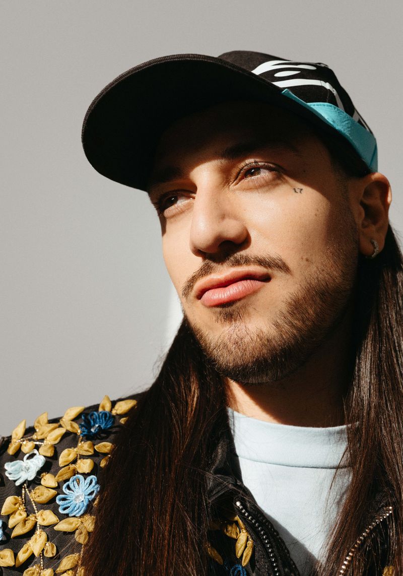 Close-up of a man with long hair, facial tattoo, and patterned jacket wearing a black and blue baseball cap against a gray background.