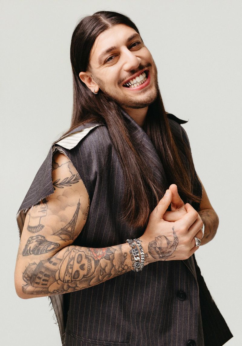 Stylish man with long hair, tattoos, and a pinstripe vest smiles confidently in a studio portrait with a neutral background.