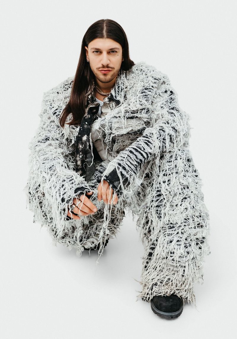 Adult male in shaggy, textured jacket and pants crouching indoors against a white background, looking toward the camera.