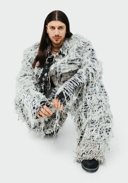 Adult male in shaggy, textured jacket and pants crouching indoors against a white background, looking toward the camera.