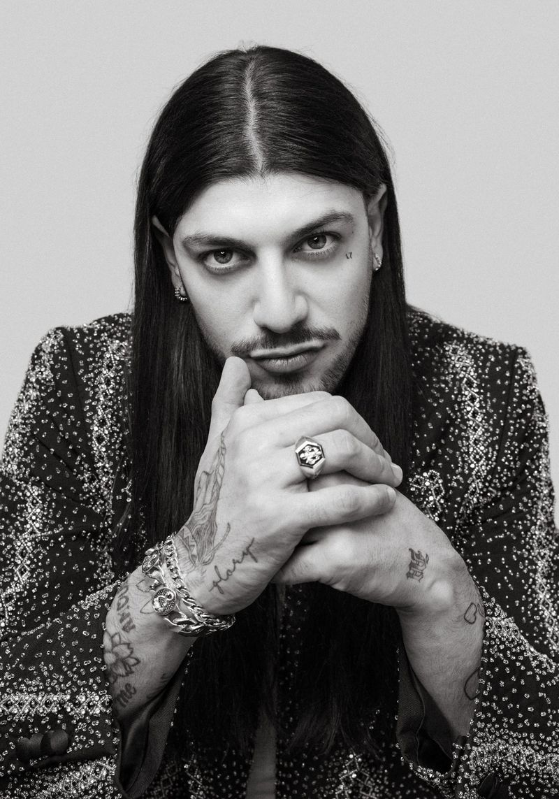 Black and white portrait of an adult male with long dark hair and tattoos, wearing a patterned blazer, posing indoors with hands clasped.