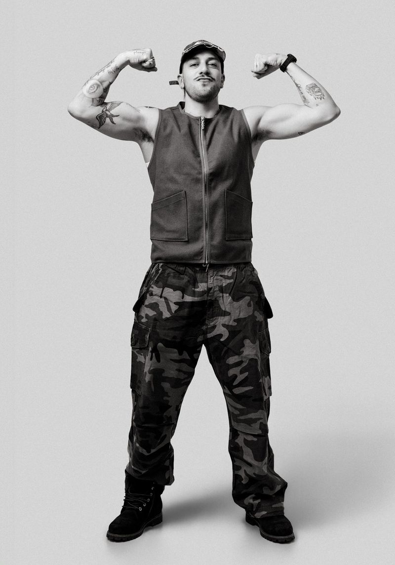 Monochrome studio shot of muscular adult male flexing in camo pants and vest. Shows confidence in a neutral gray setting.
