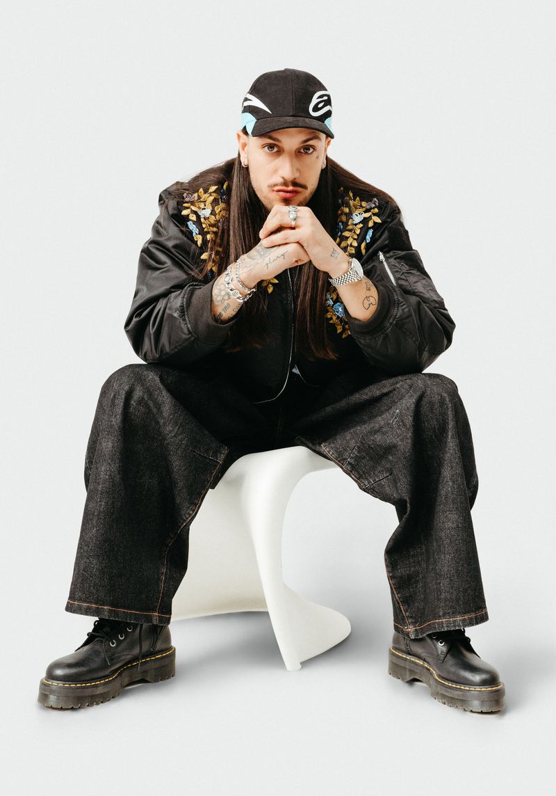 Man with long brown hair and a mustache sits on a white chair wearing a black cap, jacket and pants in a white photo studio.
