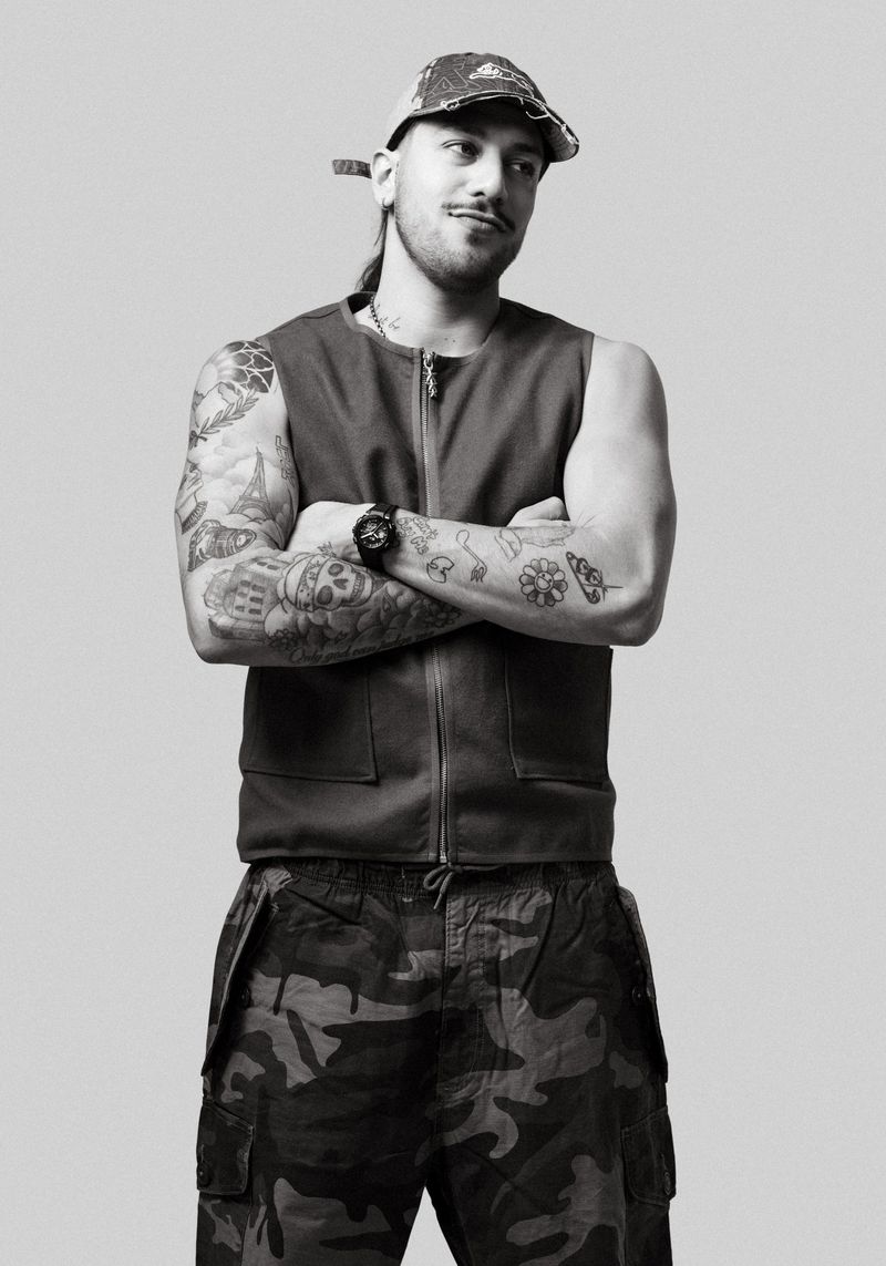 Monochrome portrait of a man with tattoos, wearing a vest and camouflage pants, arms crossed, posing in front of a neutral gray backdrop.