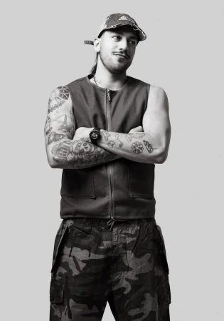 Monochrome portrait of a man with tattoos, wearing a vest and camouflage pants, arms crossed, posing in front of a neutral gray backdrop.