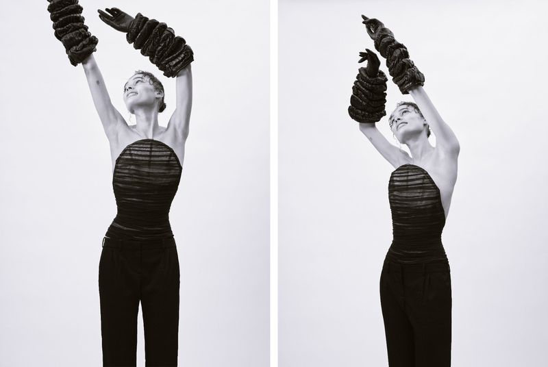 A person in a black strapless top and pants poses with gloved hands raised in a studio with a stark white background.