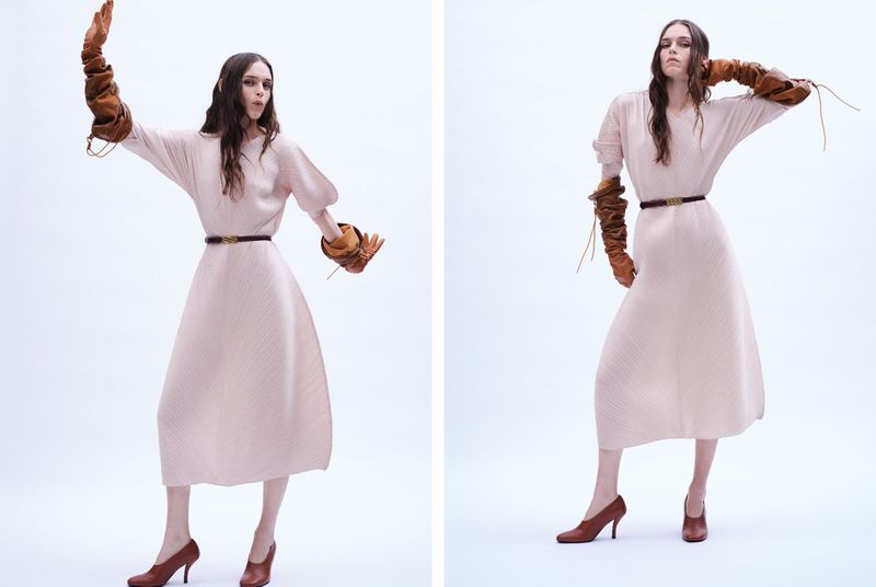 A woman in a light pink dress with long brown leather gloves and brown high heels poses in a bright white studio for a fashion shot.