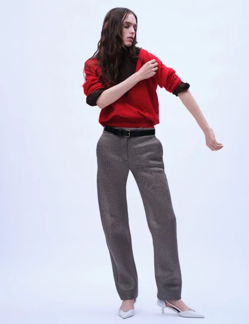 A woman in a red sweater and patterned trousers poses in front of a white background, showcasing a minimalist fashion look.