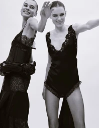 Two smiling models in black lace slip dresses posing against a plain white background in a Dazed Magazine fashion editorial.