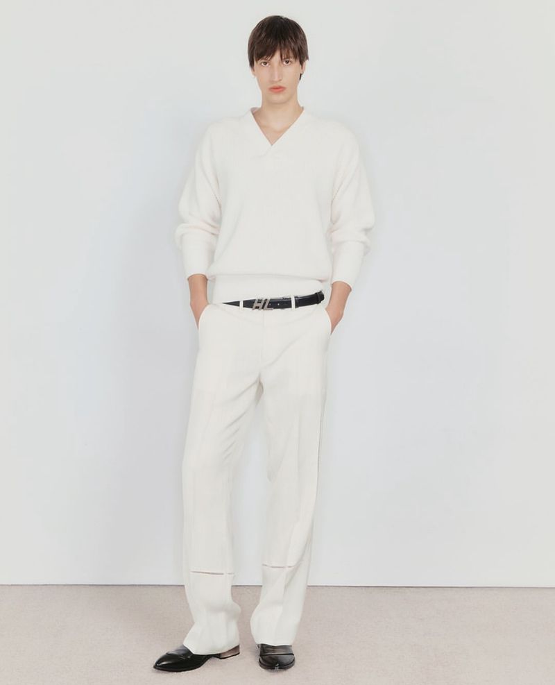 Full-length studio shot of a male model posing in an off-white V-neck sweater and trousers, with black leather belt and dress shoes.
