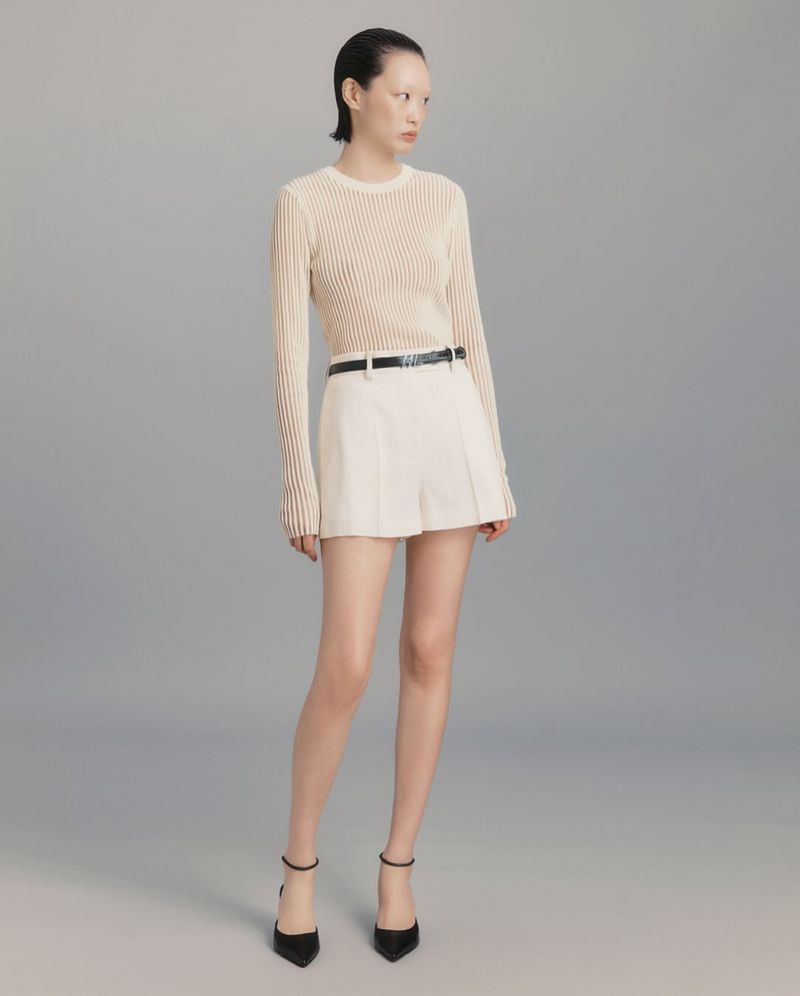 Full shot of a fashionable adult woman wearing a ribbed shirt, shorts, belt, and heels, posing in a studio with a neutral background.