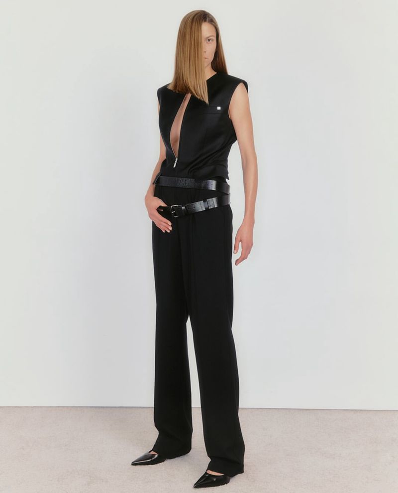 A slender woman with long blonde hair poses in a sleeveless black top, layered belts, and wide-leg trousers against a neutral backdrop.