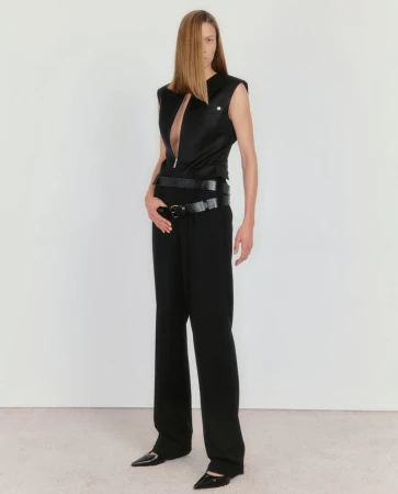 A slender woman with long blonde hair poses in a sleeveless black top, layered belts, and wide-leg trousers against a neutral backdrop.