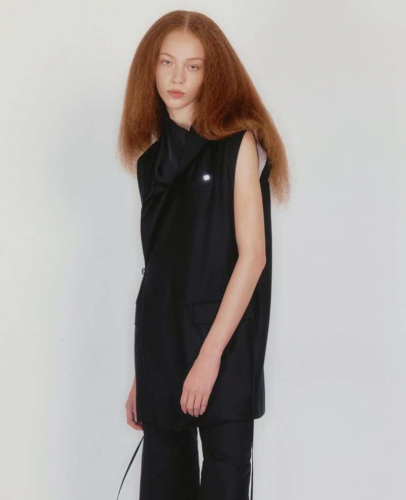 Fashion model in a black sleeveless suit with auburn crimped hair poses against a white backdrop in a studio setting.