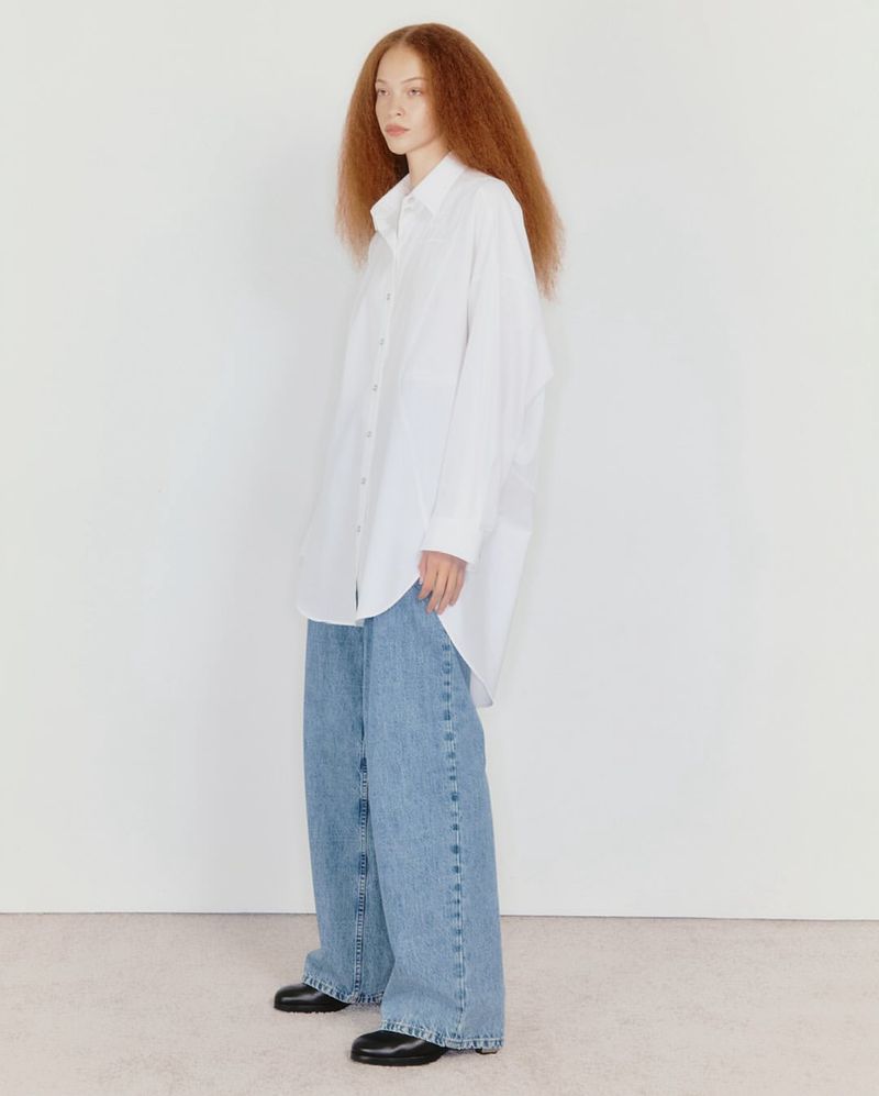 A woman with red hair in a white button-down shirt and baggy jeans posing against a white background in a fashion shot.