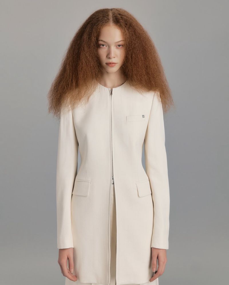 Young woman with voluminous afro hair wears a minimalist white jacket with a zipper closure in a clean, neutral studio portrait.