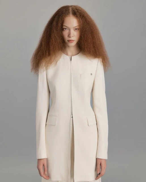 Young woman with voluminous afro hair wears a minimalist white jacket with a zipper closure in a clean, neutral studio portrait.
