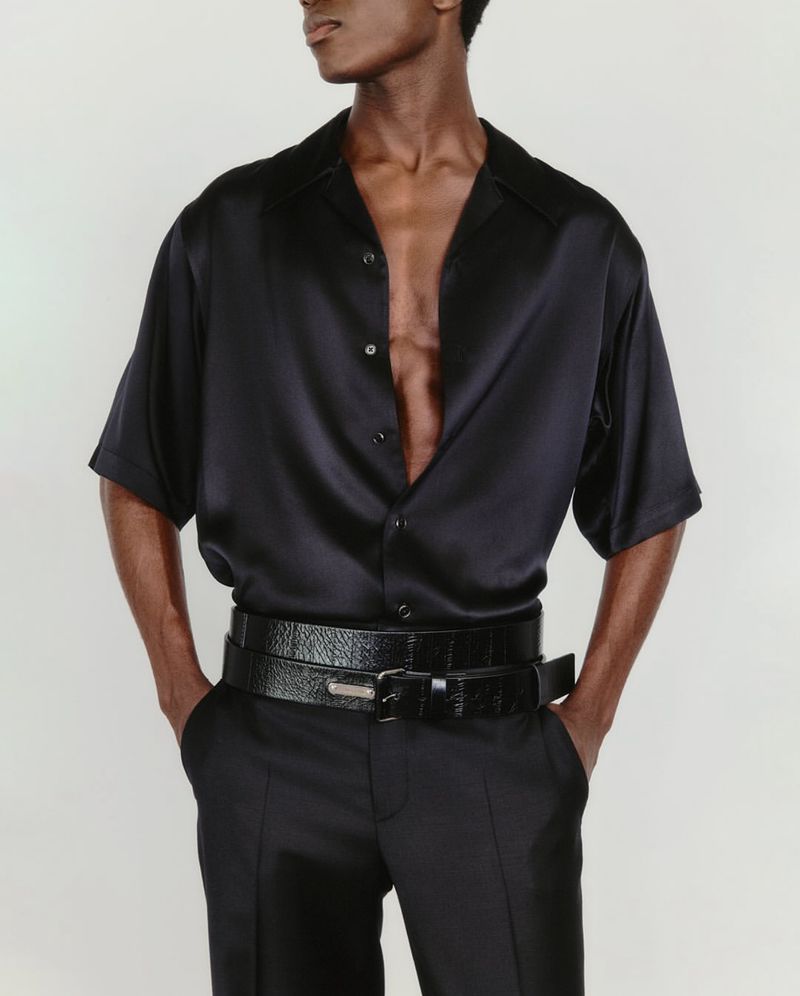 An adult male poses indoors wearing a black satin shirt with black pants and a matching belt against a neutral studio backdrop.