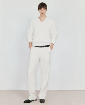Model standing against a plain white background wearing a cream V-neck sweater and matching trousers.