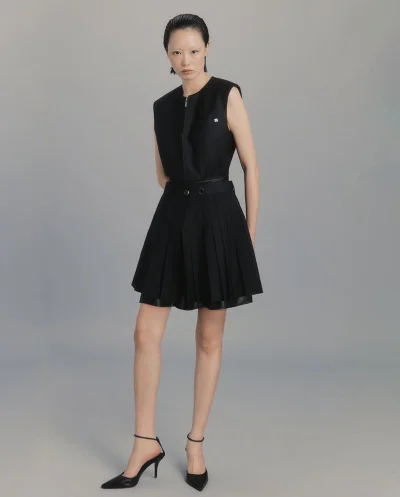 Model in a black sleeveless top and pleated skirt against a grey background for Helmut Lang Pre-Fall 2025 lookbook.