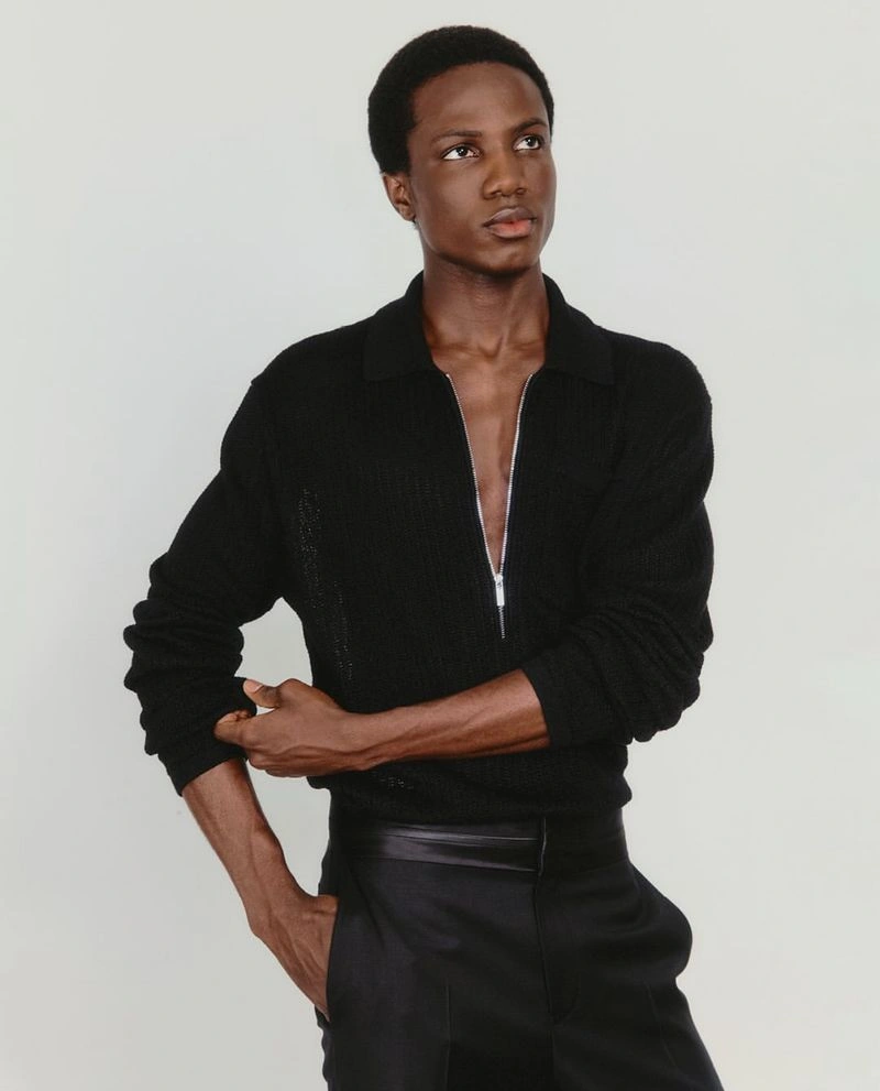 Black man wearing a black collared sweater with a zipped neckline and black trousers against a neutral background