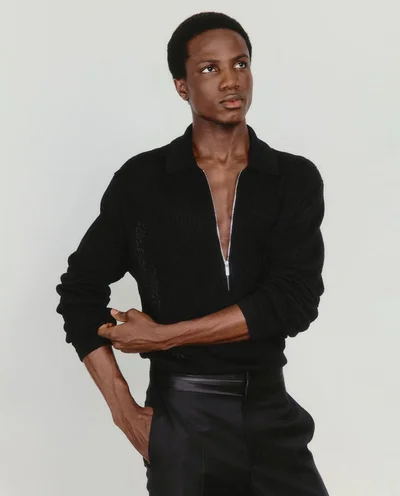 Black man wearing a black collared sweater with a zipped neckline and black trousers against a neutral background