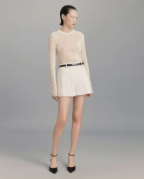 Model in ribbed cream sweater and white shorts with black belt standing against a seamless grey studio background