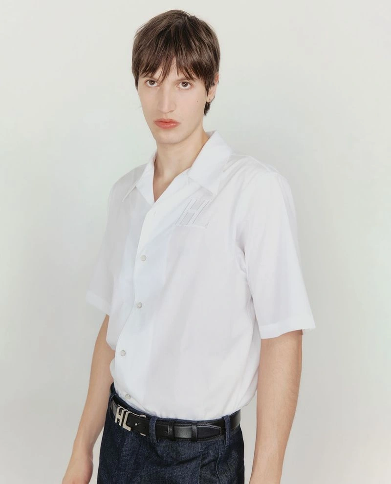 Man wearing a white short-sleeve button-down shirt and dark pants standing against a white seamless backdrop.
