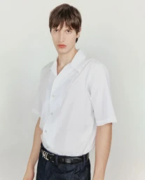 Man wearing a white short-sleeve button-down shirt and dark pants standing against a white seamless backdrop.
