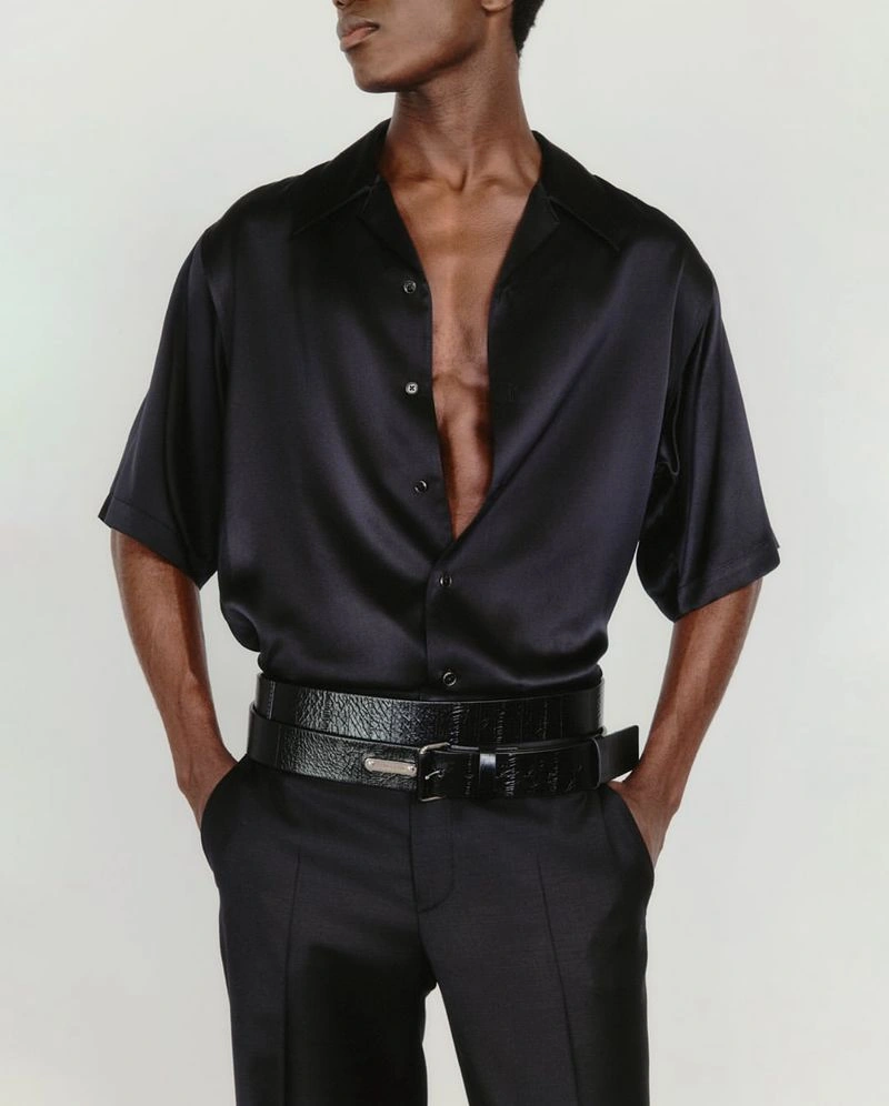 Man wearing a black silk button-down shirt and black trousers with layered belts against a white background