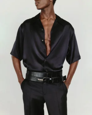 Man wearing a black silk button-down shirt and black trousers with layered belts against a white background