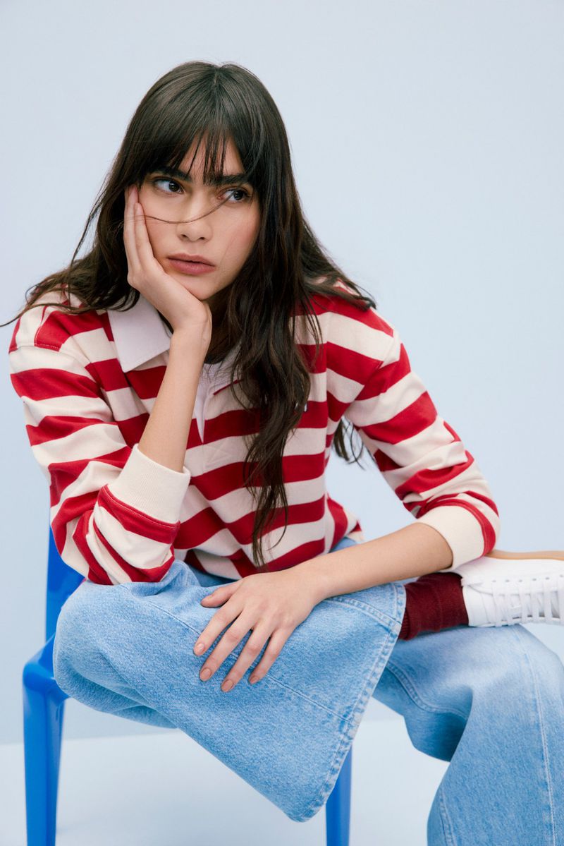 A young woman with dark hair sits on a blue chair in a red-and-white striped shirt and light-wash denim pants against a light blue backdrop.