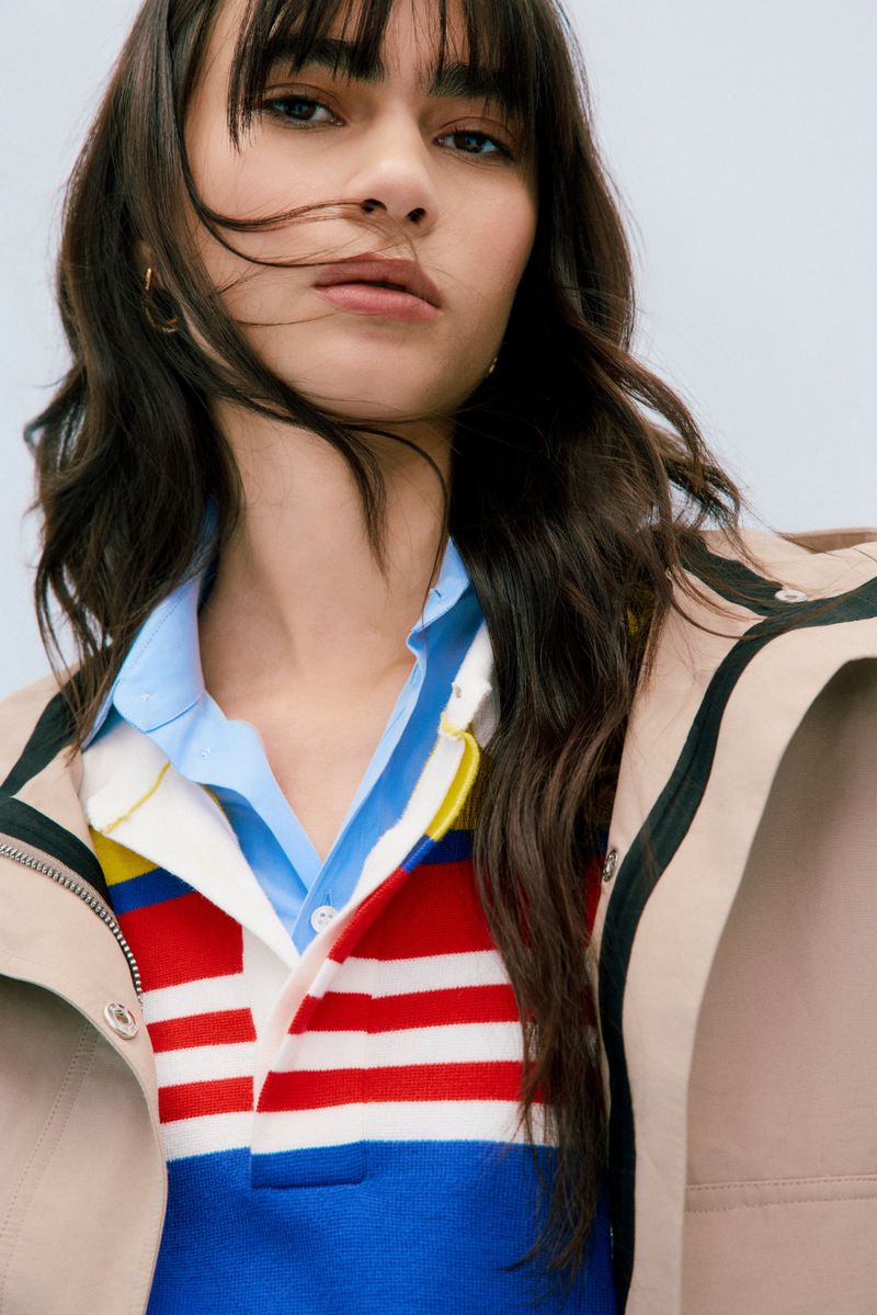Close-up of a woman with bangs wearing a red, white, and blue striped shirt and a light-brown jacket, looking directly at the camera.
