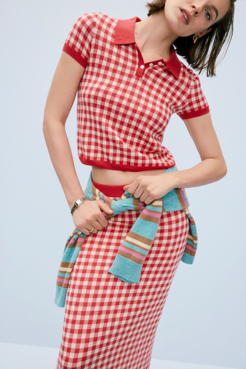 Fashion shot of a woman in a red and white gingham knit polo shirt and skirt, accessorized with a turquoise sweater, set against a light blue backdrop.