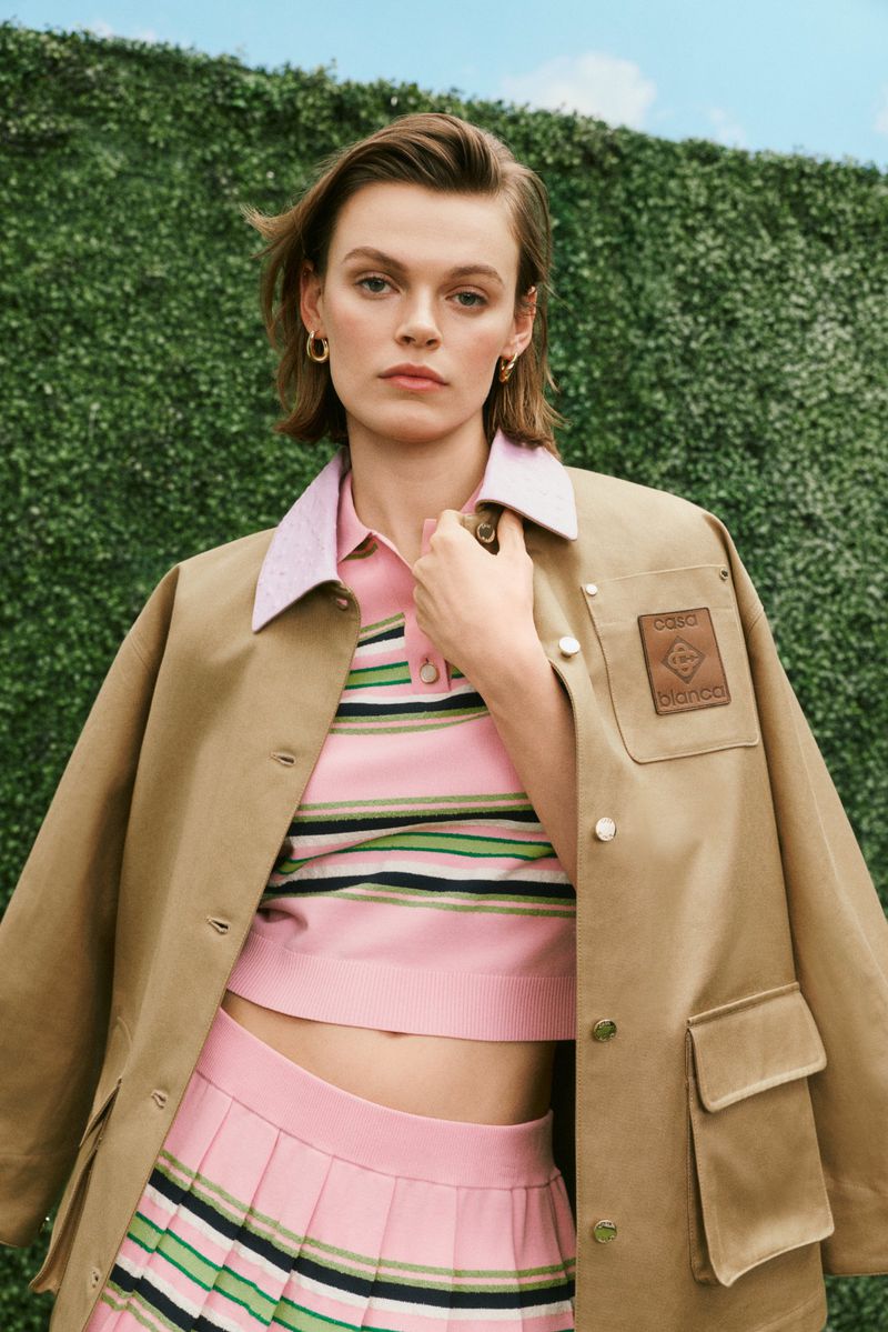 Adult female model in a pink skirt and striped crop top, wearing a tan jacket in front of a green hedge and light blue sky.