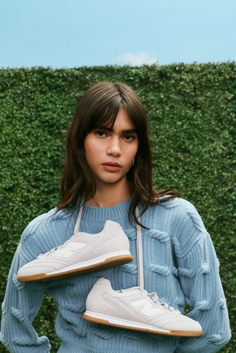 A woman with bangs and shoulder-length wavy hair wears a blue sweater, draped with beige and white sneakers against a green wall.