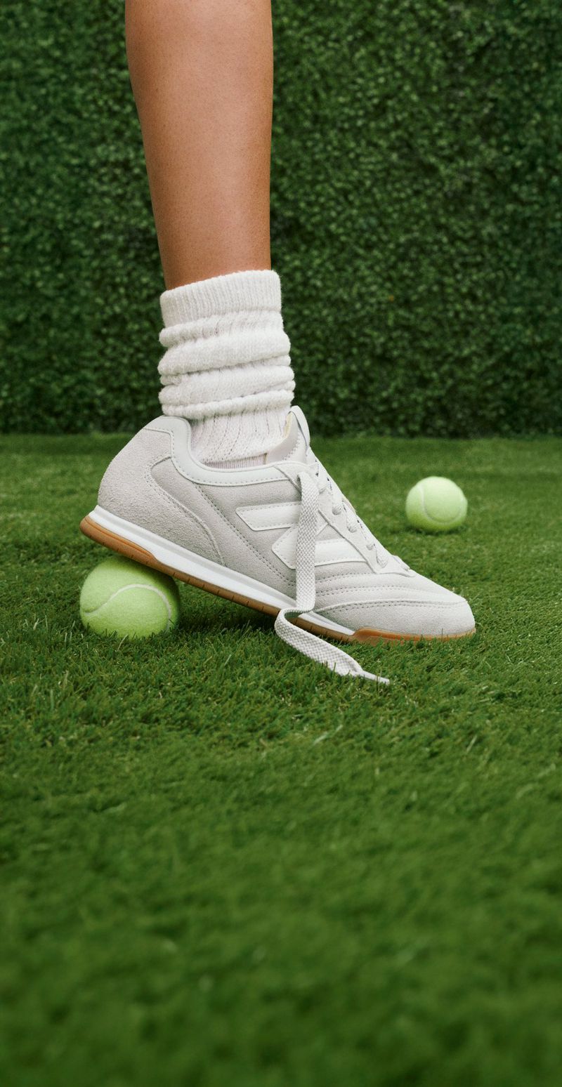 Close-up shot of a foot wearing a sneaker and a scrunched sock stepping on a tennis ball outdoors on a green grass surface.