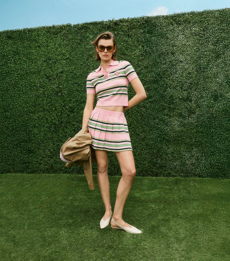 Fashion model poses in a pink and green striped tennis outfit with a jacket and white shoes against a green hedge background outdoors.