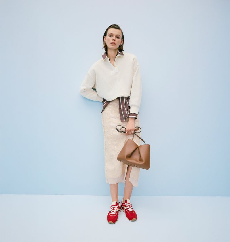 Fashion model in a cream top and skirt, accessorized with a brown bag and red sneakers, poses against a light blue backdrop.