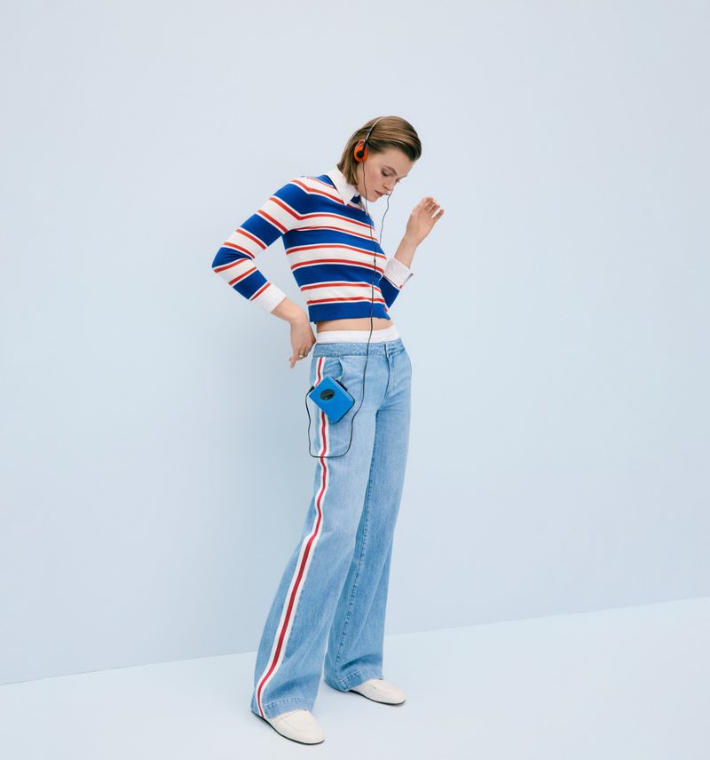 Woman in striped sweater and jeans with side stripe, wearing headphones and a portable music player, posing against light blue background.