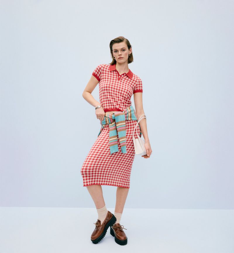 A woman poses in a red and white gingham knit midi skirt and short sleeve polo with a striped sweater tied around her waist.