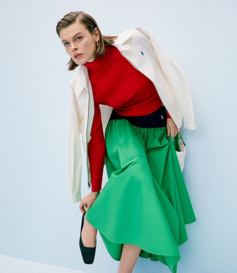 Woman in a red turtleneck, white jacket, and green skirt posing against a light blue background in a fashion shot. Full-body studio shot.