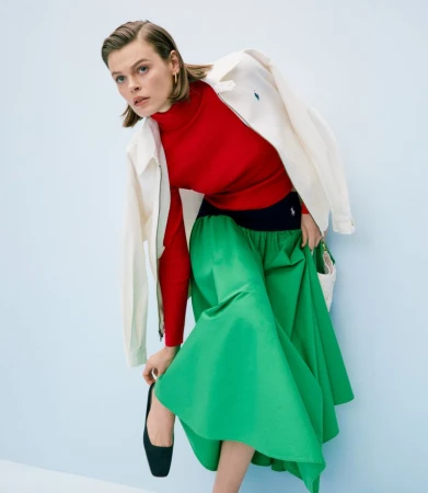 Woman in a red turtleneck, white jacket, and green skirt posing against a light blue background in a fashion shot. Full-body studio shot.
