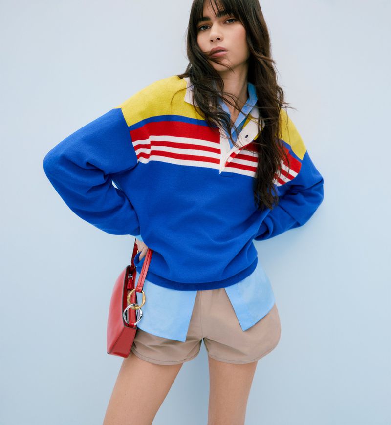 Fashion portrait of a young adult woman with dark hair, wearing a yellow, red, and blue color-blocked sweater and carrying a red purse.