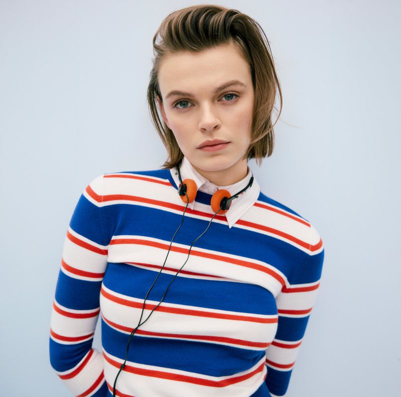 Woman in striped sweater and orange headphones gazes directly at the camera in a studio environment with a light blue backdrop.
