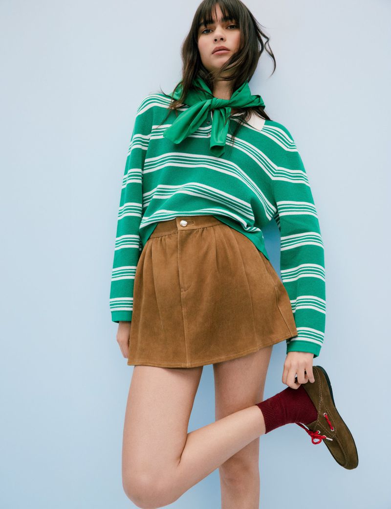 Woman poses in a green and white striped sweater with brown shorts, red socks, and brown shoes against a blue wall in studio lighting.