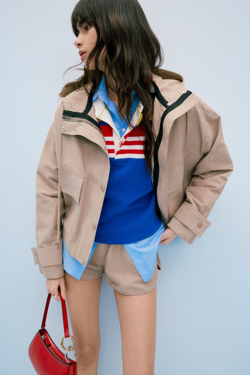 Fashionable woman in a beige jacket, blue striped shirt, and shorts, holding a red purse against a light blue background in a studio shot.