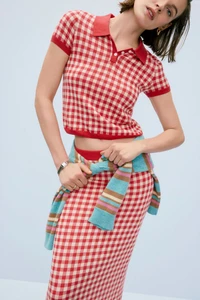 Model in a red and white gingham polo shirt and matching skirt with a striped sweater tied around her waist against a pale background