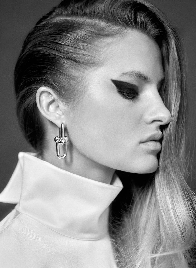 Black and white portrait of a young woman with striking black eyeliner and modern silver earrings in a high-collared white blouse.