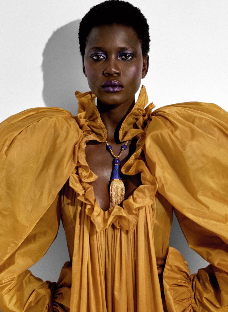 Portrait of African American woman in a gold gown with ruffle collar and statement necklace against a white backdrop in a photo studio.